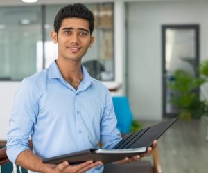 Positive Indian financial advisor holding open folder with documents and looking at camera. Smiling confident successful young businessman working in office. Business analyst concept